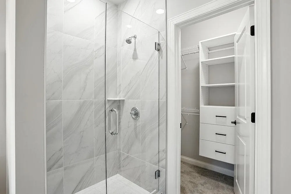 Modern bathroom interior featuring a frameless glass shower door, custom tiled shower with chrome bath accessories, and an attached walk-in closet with white melamine storage drawers and wire shelving.